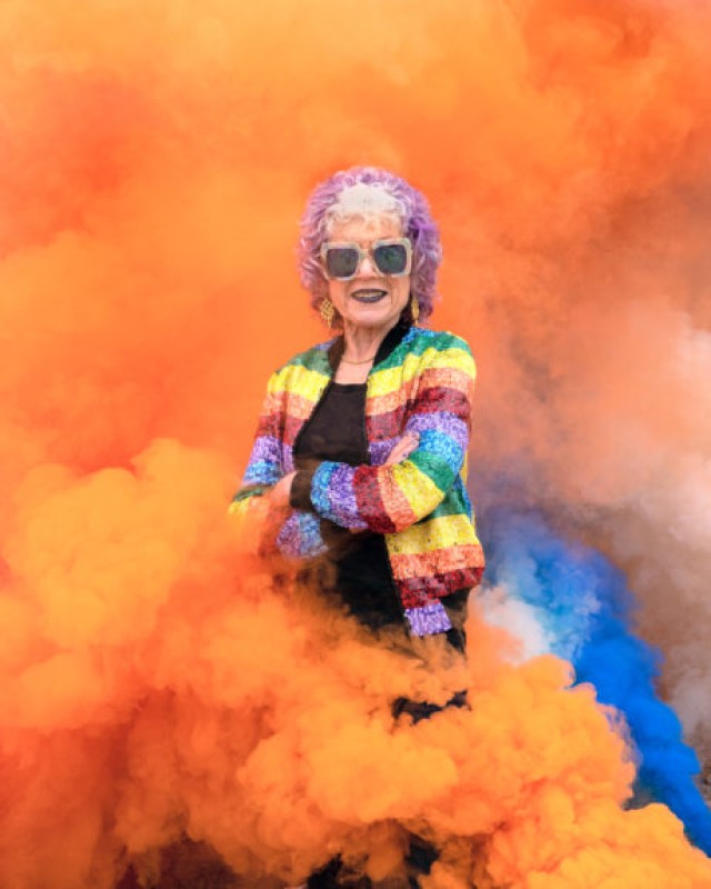 Photo of Artist Judy Chicago standing with arms crossed in a cloud of orange smoke, wearing a rainbow striped jacket, black tshirt and squarish frame sunglasses