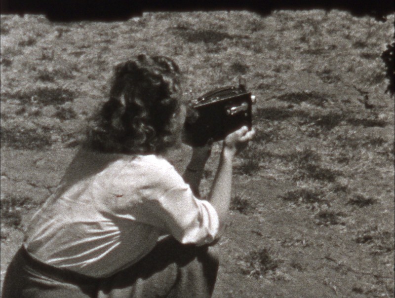 black and white film still of a 1906s era woman facing away from the camera while holding an 8 mm film camera, she has shoulder length wavy hair, wearing a white shirt and slacks, crouching while filming a subject out of frame 