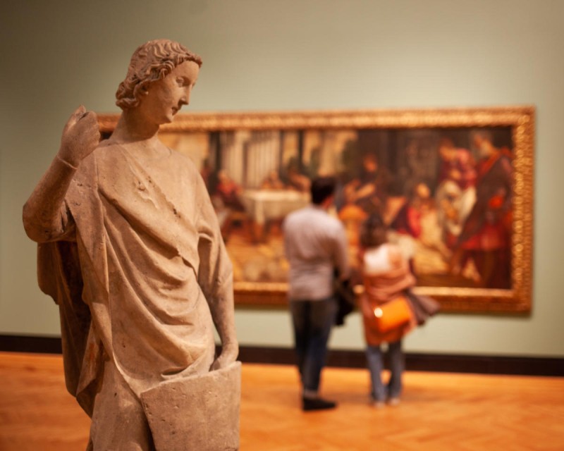 A stone statue of a woman is seen in the gallery; visitors look at art on the wall in the background.