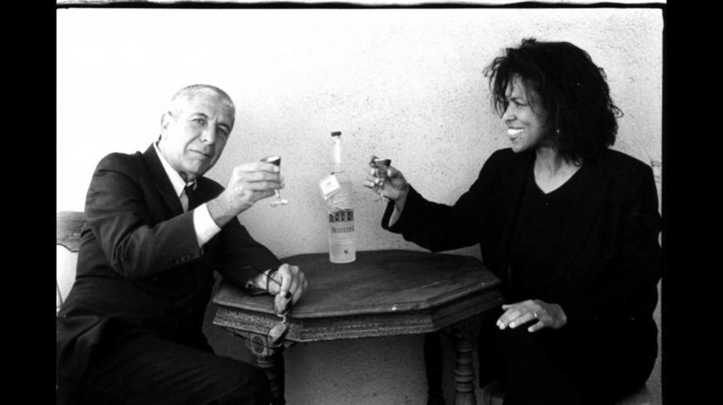Sharon Robinson and Leonard Cohen having a drink at a small wooden table. 