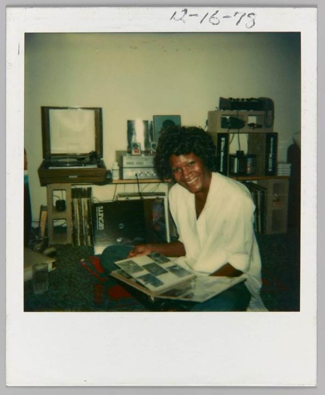 polariod photo of a Black woman sitting with a photo album open on her lap, she sits on the floor in front of various audio equipment. She is smiling at the camera. 