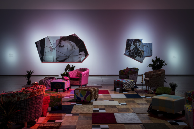 An image of a living room scene with furniture lit with pink lights. There's a carpet on the ground with living room furniture arrayed on it. Two collections of collages are on the walls behind. 