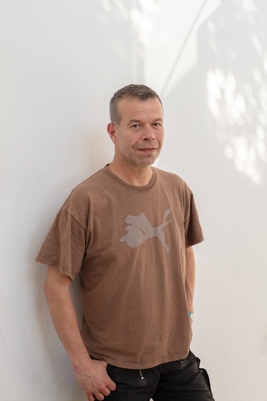 Photo of artist Wolfgang Tillmans leaning against a white wall wearing a light brown tshirt and black jeans. 