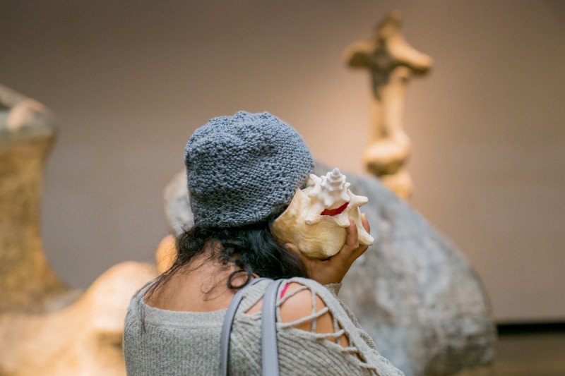 Person with long dark hair and a hat shown from behind holding a conch shell to their ear, with Henry Moore sculptures in the background.