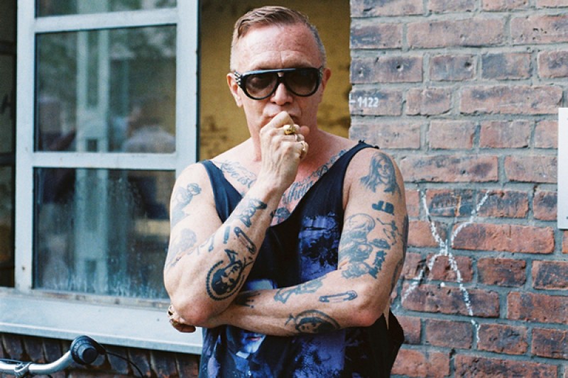 photo of Bruce LaBruce standing in front of a brick wall wearing a black tank top and dark sunglasses, arms crossed. 