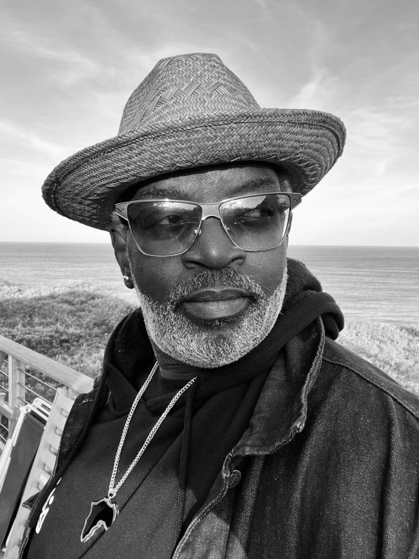 Black and white photo of Fab Five Freddy wearing a straw hat, sunglasses, hoodie, jacket and necklace with a pendant of the African continent