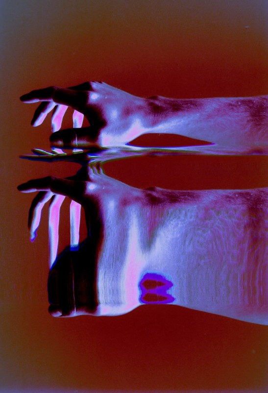 A photograph of morphed hands above liquid. The hands are bathed in blue light, on a red background seamless with the liquid. 