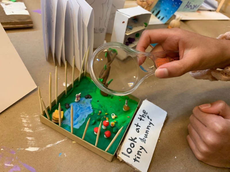 A hand holding a magnifying class over a miniature plasticine sculpture of a garden