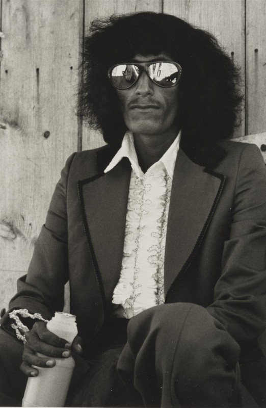 Black and white photograph of a man with shoulder-length hair, wearing reflective sunglasses, ruffled white shirt and suit with wide lapels, 