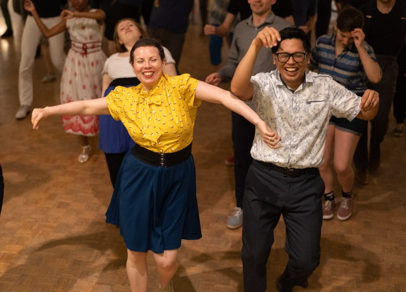 Two dancers, mid dance, a woman on the left in a bright yellow shirt and a blue dress. Person on the right, with a white collared shirt and dark blue pants. 