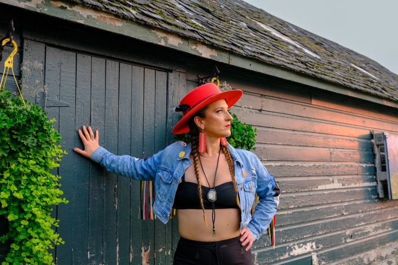 The artist leaning against a black wooden building. They 