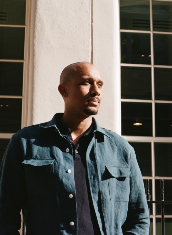 A headshot of Thomas J Price. They are looking to their left, towards sunlight. They are wearing a long sleeved button up dark green shirt, overtop of a black shirt. They are standing in front of a white building with square windows. 