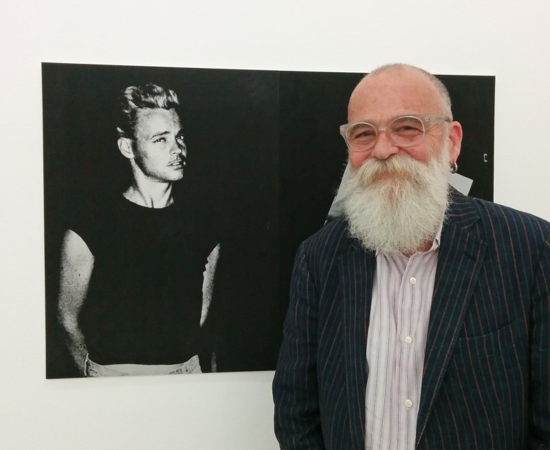 AA Bronson smiling and standing in front of a photo of their youth. They have a long beard and are wearing a dark coloured jacket.