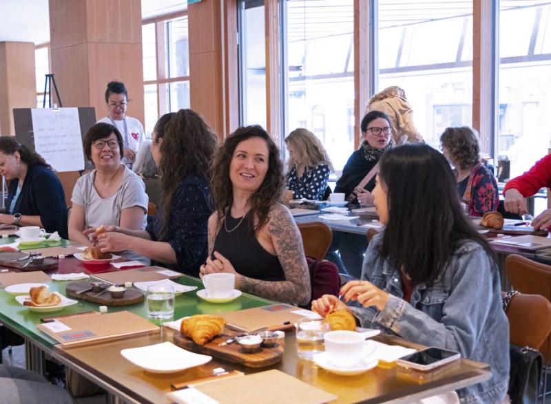 Group of people seated at a table with coffee and pastries, smiling in a bright room.