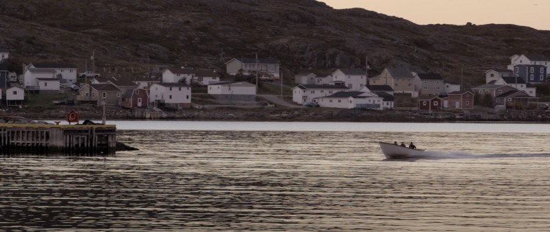 Still image from the film Hand Line Cod, showing a fishing boat on the ocean with an oceanside town in the background