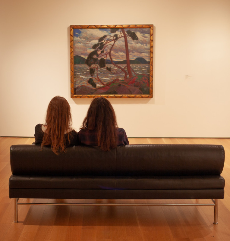 Two visitors seated on a couch with their backs to the camera. Together they are looking at Tom Thomson's "The West Wind" (1916–1917), hanging on the wall opposite of them.