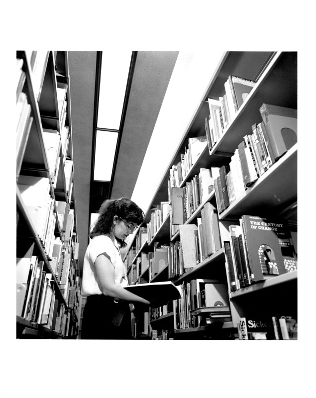 Black and white photo of young white woman with long hair and glasses reading a book between library shelves. From the 1980s